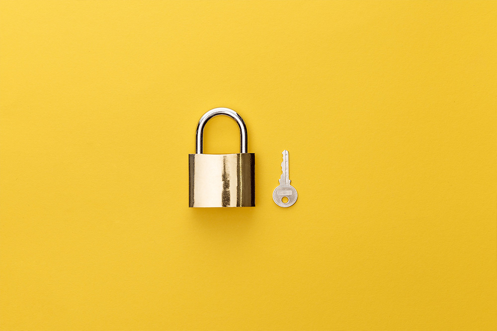 A small and shiny gold padlock lies on a bright yellow background, with a small silver key lying next to it.